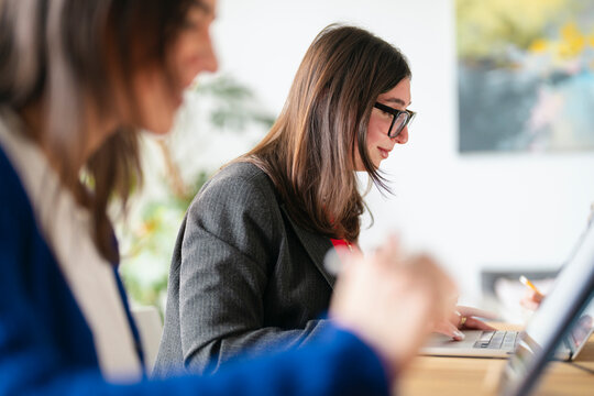 Two women collaborate at a desk, one typing on a laptop while the other writes, focused on their tasks in a modern office setting.