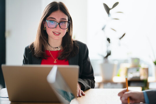 A young woman wearing glasses works on her laptop while a colleague takes notes at a desk in a bright office setting.