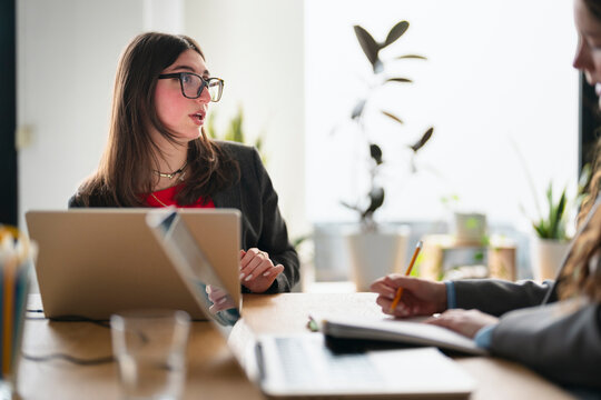 Two women in a professional setting discuss business, one speaking while the other takes notes on a document with a pencil.