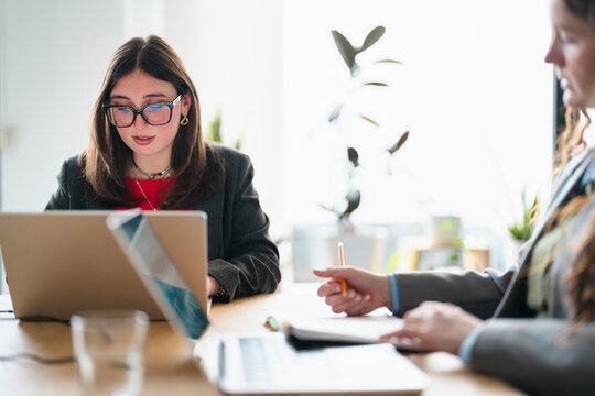 Two women in business attire collaborate at a desk, one using a laptop and the other taking notes with a pencil.