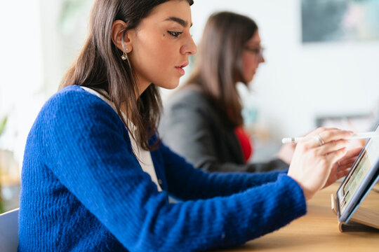 A young woman in a blue sweater uses a stylus to interact with a tablet computer, with another person blurred in the background.