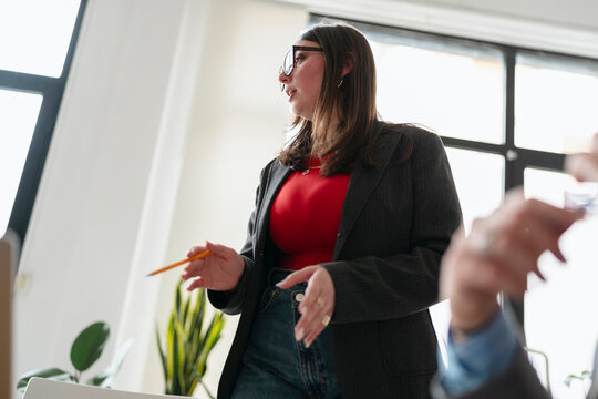 A woman in a blazer and jeans holds a pencil while speaking to colleagues in a bright office setting.