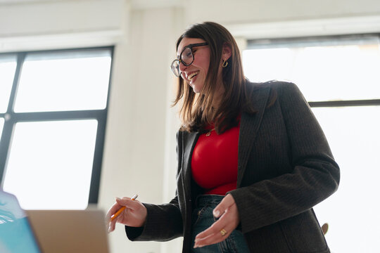 A young woman in glasses and a blazer smiles while holding a pencil and looking at a laptop.