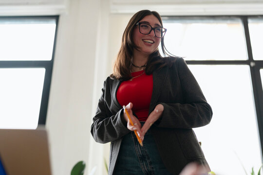 A young woman wearing glasses and a blazer smiles while holding a pencil, suggesting she is engaged in a creative or educational activity.