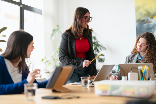 Three women collaborate in a modern office setting, engaged in discussion and working on laptops and tablets, fostering a productive team environment.