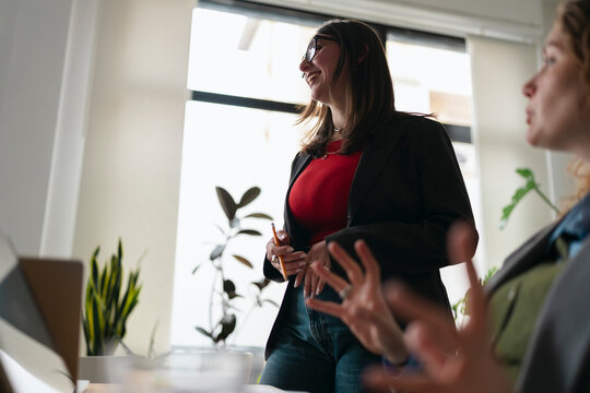 A woman in glasses and a blazer smiles while holding a pencil, presenting an idea to colleagues in a bright office setting.