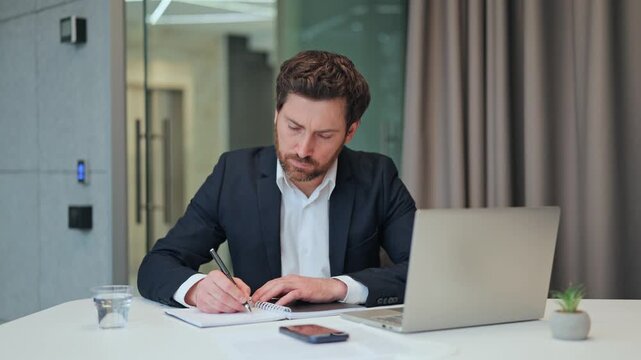 Businessman working at desk, concentrating on tasks. Focused individual notes information from laptop into notebook, reflecting serious dedication to work in modern office, commitment effort.
