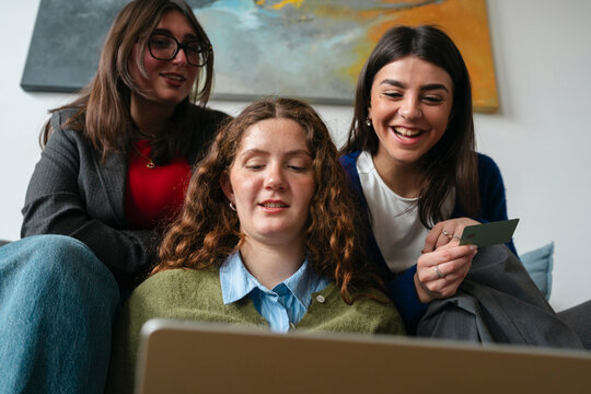 Three young women are gathered around a laptop, one holding a card, as they engage in online activity together.