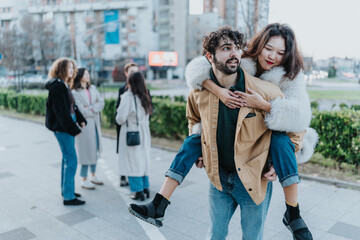 Fototapeta premium A man carries a woman on his back as they walk with friends in a city park, showing playful affection.