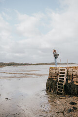 Obraz premium A woman stands on a ladder overlooking a body of water. The sky is cloudy, and the water is calm. The scene is peaceful and serene, with the woman enjoying the view and the quiet atmosphere