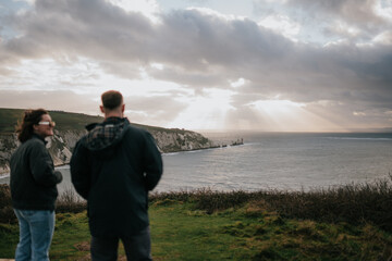 Fototapeta premium Two people standing on a grassy hill overlooking the ocean. The sky is cloudy and the sun is shining through the clouds