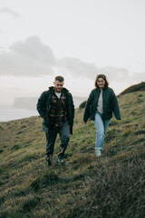 Fototapeta premium A man and woman are walking on a hillside. The man is wearing a plaid jacket and the woman is wearing a green jacket. The sky is cloudy and the couple is enjoying the view of the ocean