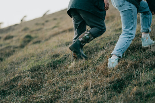 Two people are running up a hill in the grass. The man is wearing a black jacket and the woman is wearing blue jeans