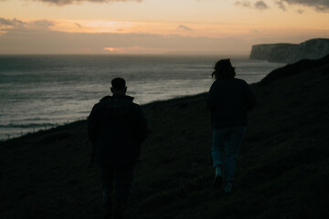 Two people walking on a hill overlooking the ocean. The sky is orange and the water is calm