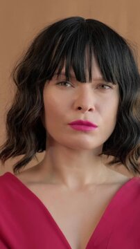 White Woman Casting Director Evaluating Audition With Skeptical Expression And Poised Posture In Red Dress, Neutral Brown Studio Background, Pink Lipstick, Bangs Framing Face, Fashion Confidence