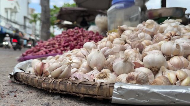 Fresh garlic bulbs are piled on a wooden pallet placed on the ground, with open street surroundings and active market environment visible around the stall. Street food market.