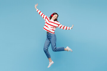 Full body young woman she wear red striped sweatshirt casual clothes jump high listen to music in headphones with outstretched hands isolated on plain pastel light blue background. Lifestyle concept. © ViDi Studio
