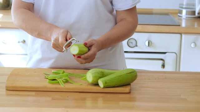 Woman is cutting a cucumber on a wooden cutting board. She is using a mandolin to slice the cucumber