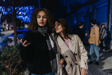 Fototapeta premium Two stylish women pose on a city sidewalk at night, one holding a phone. They lean together under blue decorative lights, dressed in coats, looking relaxed.