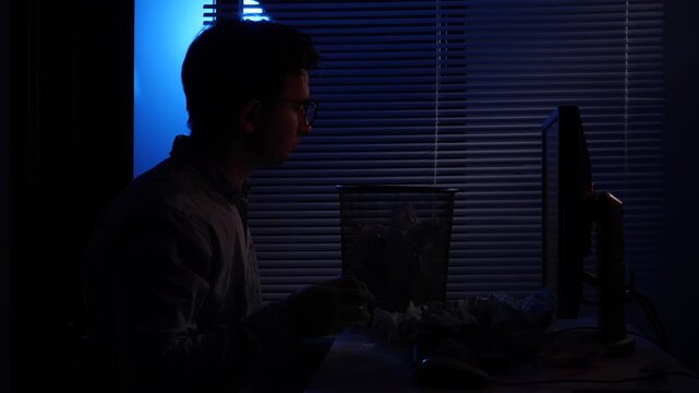 Man working late in a dark office, exhausted and frustrated, crumpling paper and angrily tossing it into a wastebasket beside his computer during a tense deadline push