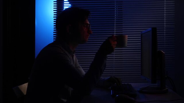 Young man focused at his computer, typing and sipping coffee in a dark room lit by blue evening light through blinds, suggesting late night overtime and concentration