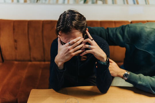 A man in a dark hoodie sits on a brown sofa with his hands over his face. A nearby friend reaches out, suggesting support in a casual living room scene.