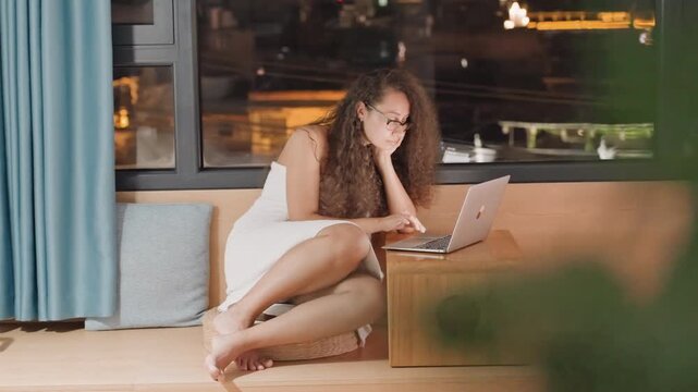 Woman in towel working laptop inside quiet hotel room near window with city lights, wooden stool as table, barefoot and focused typing, cozy curtain and warm lamp glow, digital nomad editing copy,