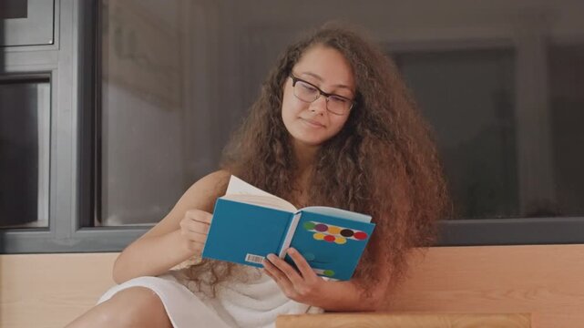 Woman reading in hotel window seat, turning blue book pages with intent, warm wood bench and dark window background, latenight solitude and creative reflection, gentle gestures suggesting writer