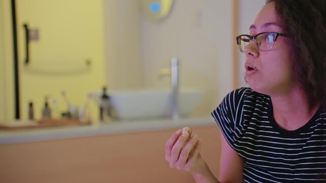 Woman eating cookie in hotel room by bathroom vanity, mirror reflection and warm yellow light, taking a bite with smile, glasses and curly hair, candid latenight relaxation and playful mood