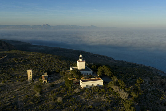 Aerial view of the stark white lighthouse standing sentinel against the hazy horizon, where sea meets sky in a soft, ethereal embrace, Santa Pola, Comunidad Valenciana, Spain.