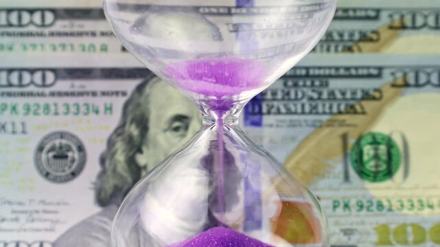 Purple sand dropping through the bulb of the hourglass and dollar money on the background. Hourglass with sand stand among money. Deadline, business planning, finance and time is money concept.