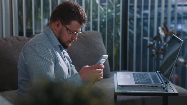 Man replying to message on smartphone while working on laptop sitting on sofa and briefly interrupting computer work
