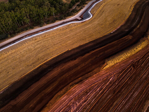 Aerial view of contrasting rust-red earth meeting golden fields near a dense green forest, a stark boundary defined by a stark white road, Aluminium Refinery, Western Australia, Australia.