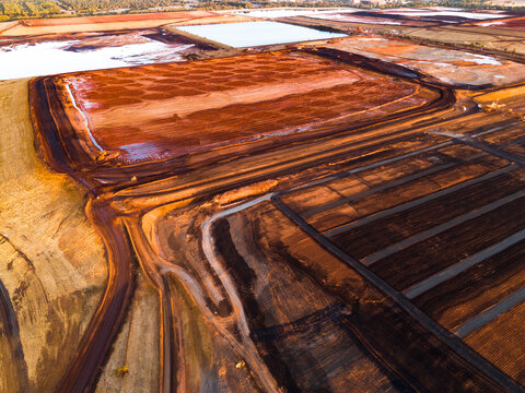 Aerial view of rust-colored earth meets stark white pools under a hazy sky, the refinery's vast landscape etched with geometric precision, Aluminium Refinery, Western Australia, Australia.