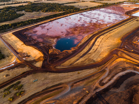 Aerial view of a stark contrast between the red-tinted water and the surrounding golden fields, creating a surreal landscape, Aluminium Refinery, Western Australia, Australia.