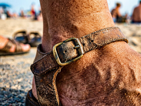 Photographie en tr&egrave;s gros plan d'une cheville humaine avec une sandale en cuir us&eacute; et une boucle m&eacute;tallique sur une plage ensoleill&eacute;e