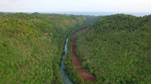 Aerial drone footage of a hidden forest and mountains valley, with the touristic Maron river, in Pacitan regency, Java island, Indonesia, and many rounded hills in the background, at sunset