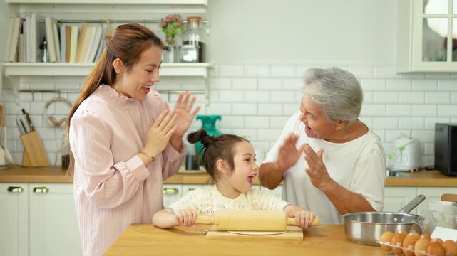 A happy multi generational Asian family, grandmother, mother, and little daughter enjoy baking together in a bright kitchen. The child rolls dough as they cheer, capturing a warm bonding moment.