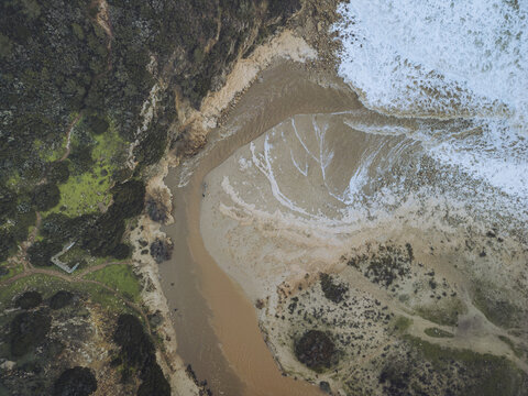 Aerial view of the rugged coastline where the river meets the tumultuous sea, creating a mesmerizing dance of sand, water, and foam, Budens, Faro, Portugal.