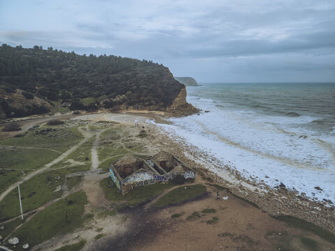 Aerial view of the Boca do Rio, where the ocean's foamy embrace meets weathered ruins beneath a brooding sky, Budens, Faro, Portugal.