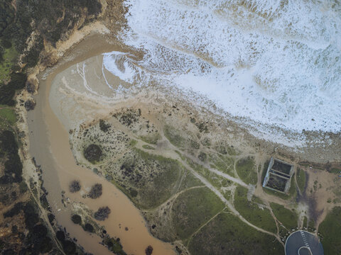 Aerial view of the confluence of a muddy river meeting the foamy ocean waves creating a striking contrast of colors and textures, Budens, Faro, Portugal.