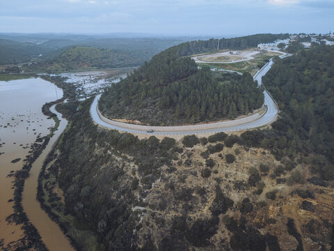 Aerial view of a winding road snaking through the verdant hills, contrasting with the muddy river and the distant structures, Boca do Rio, Budens, Faro, Portugal.