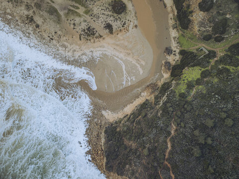 Aerial view of where crashing ocean waves meet the tranquil sands of Boca do Rio beach, divided by rugged, dark green vegetation, Budens, Faro, Portugal.