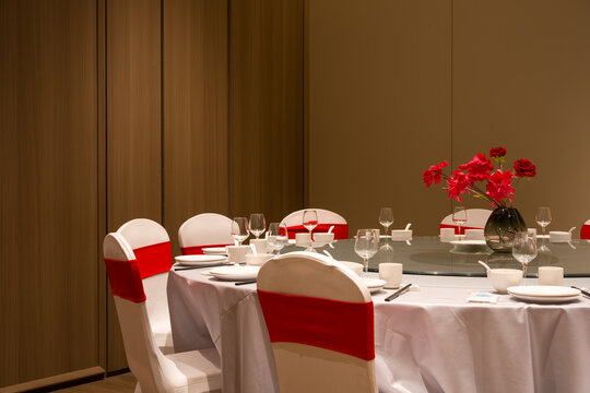 Hotel private dining room with round table set for meal featuring red flowers white chairs and elegant tableware