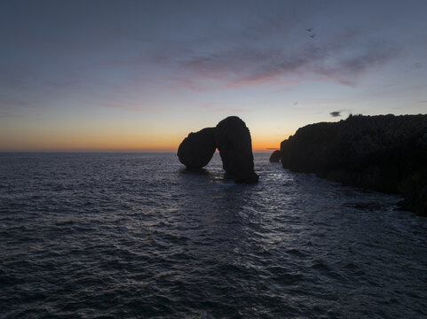 Aerial view of the dark, rocky arch emerging from the shimmering, blue sea, with the horizon ablaze in the colors of the setting sun, Llanes, Asturias, Spain.