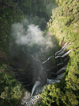 Aerial view of a cascading waterfall plunges into a verdant gorge, mist rising to meet the dense forest canopy, Java, Java, Indonesia.
