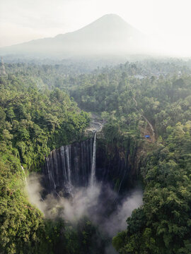 Aerial view of the majestic Tumpak Sewu waterfall cascading through lush green vegetation, with the hazy silhouette of Mount Semeru in the distance, Java, Indonesia.