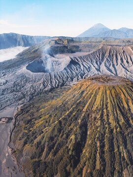 Aerial view of the rugged volcanic landscape, a symphony of greys and browns, with Mount Bromo's caldera steaming gently under a vast sky, Java, Indonesia.