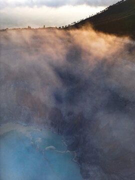 Aerial view of ethereal mists swirl above the acidic turquoise lake, contrasting with the dark, rugged terrain and distant city skyline, Java, Java, Indonesia.