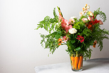 A bouquet of roses, gladioli, zinnias, and eustomas stands in water. Carrots with green tops are part of the arrangement on a plain table against a light background © pundapanda